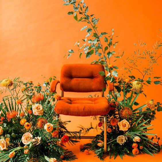 Orange chair against orange wall, surrounded by greenery and flowers.