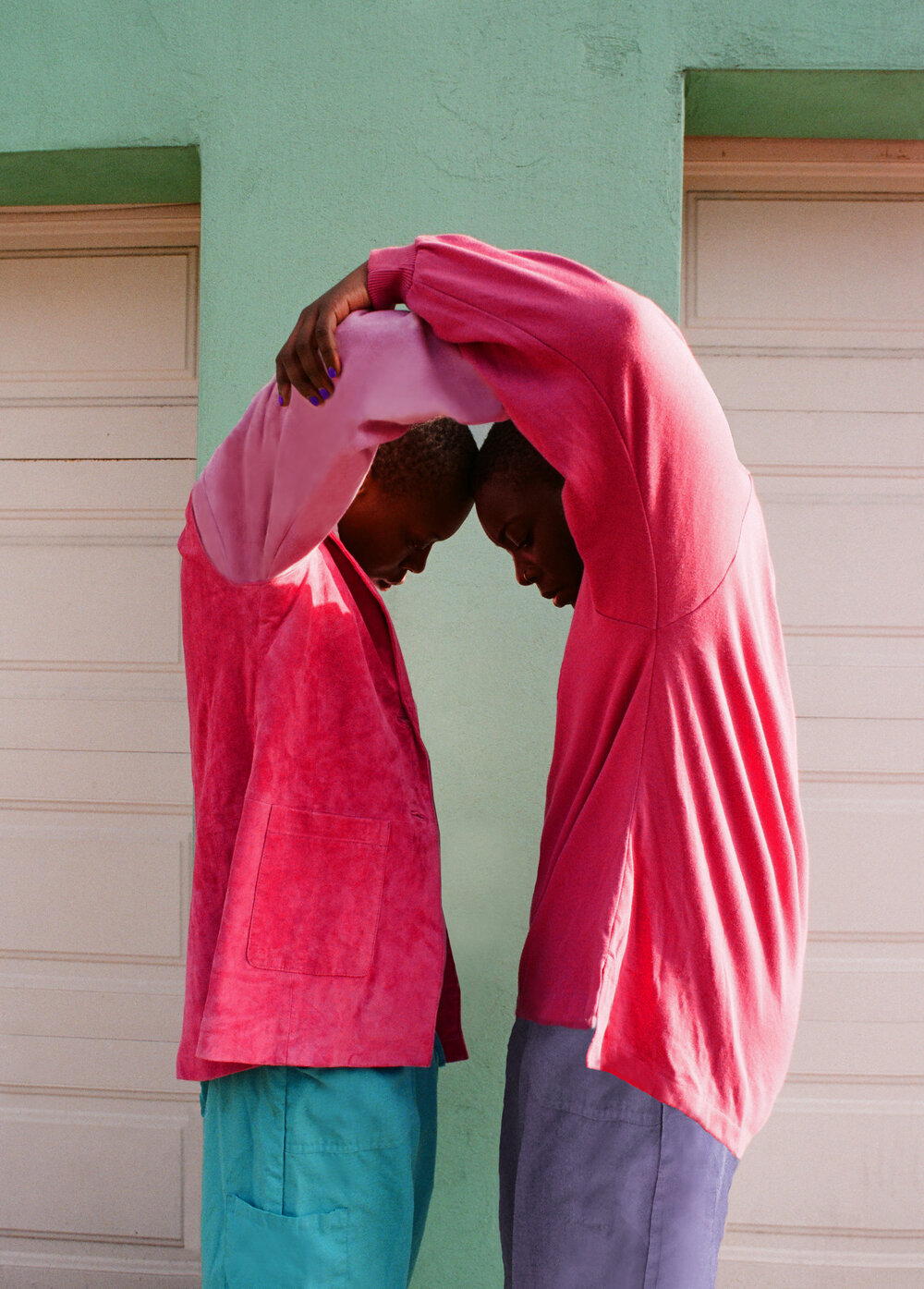 Two twin models wearing identifical clothes, mirroring one another in an over-the-head embrace.